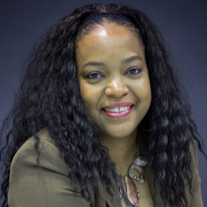 The image shows a woman with long, curly black hair and a warm smile. She is wearing a brown blazer and a necklace. The background is a plain, dark color. She appears to be looking directly at the camera.